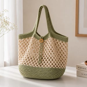 May include: A two-toned crochet tote bag featuring a solid sage green base and top trim with a cream-colored open diamond mesh body. The bag includes two matching sage green handles and a single wooden button closure at the center top opening.