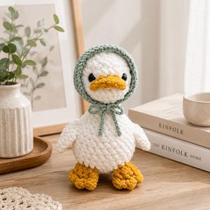 May include: A white crocheted duck toy with a yellow beak and feet. The duck wears a sage green crocheted bonnet tied under the chin with a bow. The toy has black bead eyes and is crafted from soft, textured yarn, standing upright on a flat surface.