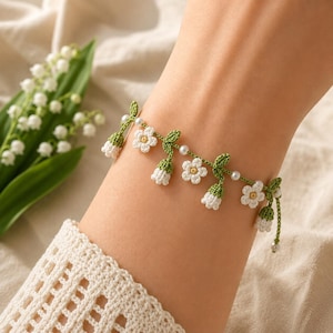May include: A delicate bracelet featuring green crocheted stems and leaves, small white five-petaled flowers with yellow centers, white bell-shaped lily of the valley blossoms, and small white spherical beads strung along the thin green cord.