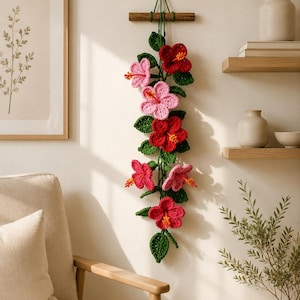 Puede incluir: Un adorno de pared vertical que presenta flores de hibisco tejidas a ganchillo en tonos rojos y rosas, acentuadas con hojas verdes tejidas. El arreglo floral está suspendido de un listón de madera horizontal mediante hilos verdes.