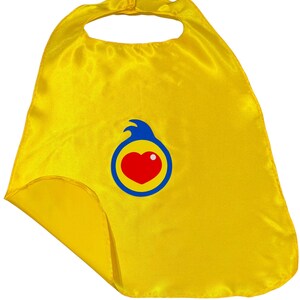 May include: A bright yellow superhero cape made of shiny fabric. The cape features a circular emblem with a red heart, a white dot, and a blue and yellow border. The cape has a neck opening and a Velcro closure.