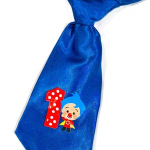 May include: A bright blue satin tie featuring a red number one with white polka dots and a cartoon character. The character has blue hair, a red nose, and a yellow cape. This tie is perfect for a child's first birthday.