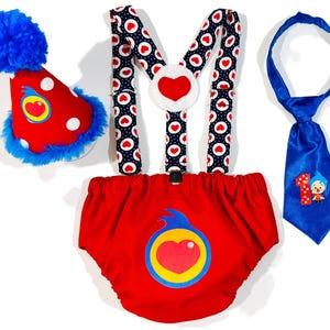 May include: A festive baby outfit set featuring a red diaper cover, suspenders, a party hat, and a tie. The diaper cover and hat have a heart design. The suspenders have a heart and polka dot pattern. The blue tie has a number 1 and a cartoon character.