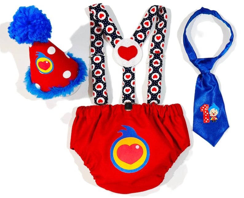 May include: A festive baby outfit set featuring a red diaper cover, suspenders, a party hat, and a tie. The diaper cover and hat have a heart design. The suspenders have a heart and polka dot pattern. The blue tie has a number 1 and a cartoon character.
