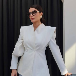 Puede incluir: Una americana blanca de doble botonadura con hombros estructurados, mangas largas y escote en V, combinada con vaqueros azul claro. La americana presenta dos botones blancos a juego en la cintura y un corte entallado, con gafas negras.