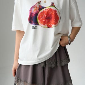 May include: A white short-sleeved t-shirt featuring a graphic of a whole fig and a sliced fig with the text FRUIT. FRAGANCIAS, paired with a grey tiered skirt featuring lace trim along the edges of each layer and a thin gold chain strap.