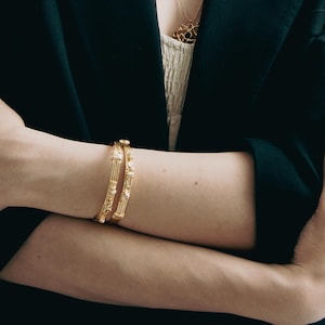 Può includere: Bracciali dorati con disegni intricati adornano un braccio, su uno sfondo di blazer nero. I braccialetti presentano motivi e trame dettagliate. È visibile anche una collana d'oro. Lo sfondo è neutro.
