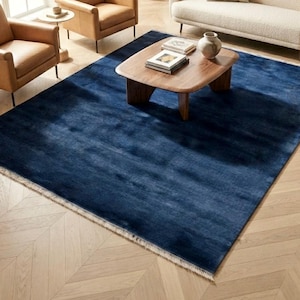 Puede incluir: Una alfombra rectangular grande de color azul marino sólido y profundo con un acabado de textura suave y sutil, con flecos cortos de color claro a lo largo de los dos bordes más cortos, colocada sobre un suelo de madera clara.