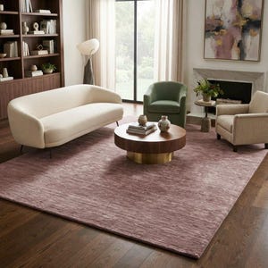 Puede incluir: Una alfombra rectangular grande en un tono malva apagado con un sutil patrón de textura horizontal. La alfombra está colocada sobre un suelo de madera oscura, sirviendo de base para un sofá color crema, un sillón verde y una mesa de centro redonda.