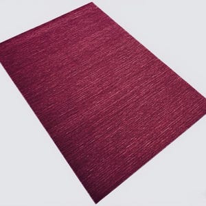 Puede incluir: Una alfombra rectangular de color magenta intenso que presenta una sutil textura lineal horizontal en toda la superficie. La alfombra se presenta desde una perspectiva en &aacute;ngulo, mostrando su color uniforme y su dise&ntilde;o de patr&oacute;n tejido consistente.