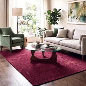 Puede incluir: Una alfombra rectangular grande de color rojo burdeos intenso con una superficie tejida y texturizada. La alfombra est&aacute; colocada sobre un suelo de madera debajo de una mesa de centro con tapa de cristal, un sof&aacute; claro y un sill&oacute;n de terciopelo verde.