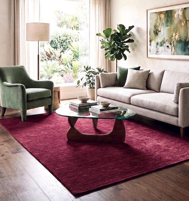 Puede incluir: Una alfombra rectangular grande de color rojo burdeos intenso con una superficie tejida y texturizada. La alfombra est&aacute; colocada sobre un suelo de madera debajo de una mesa de centro con tapa de cristal, un sof&aacute; claro y un sill&oacute;n de terciopelo verde.