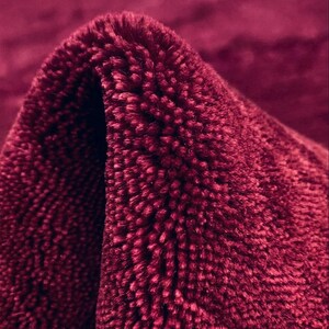 Puede incluir: Una alfombra de color burdeos intenso con una textura gruesa de pelo largo. El material presenta una superficie densa y con mechones que crea un aspecto suave y afelpado. La tela est&aacute; doblada para revelar la profundidad de las fibras rojas.