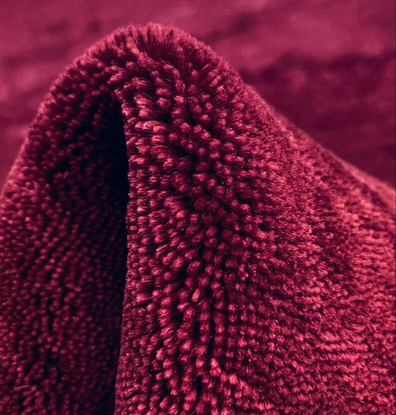 Puede incluir: Una alfombra de color burdeos intenso con una textura gruesa de pelo largo. El material presenta una superficie densa y con mechones que crea un aspecto suave y afelpado. La tela est&aacute; doblada para revelar la profundidad de las fibras rojas.