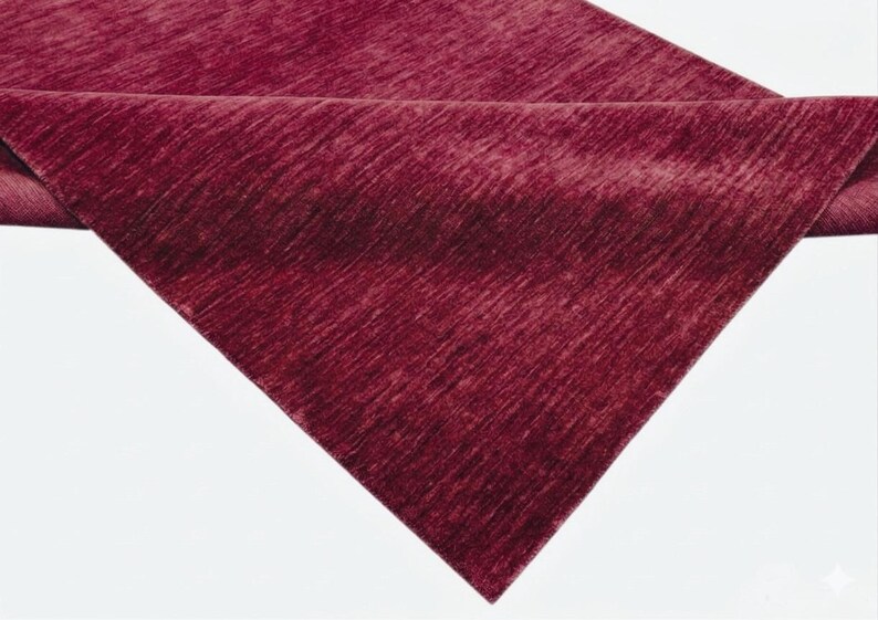 Puede incluir: Un camino de mesa de color burdeos intenso con una textura acanalada direccional sutil. La tela presenta un acabado consistente y ligeramente lustroso en toda su superficie, con una esquina doblada que revela el reverso del material.