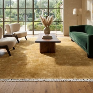 Puede incluir: Una alfombra rectangular grande de color amarillo mostaza sólido con una textura de pelo corto y detalles de flecos blancos a lo largo del borde inferior. La alfombra está colocada sobre un suelo de madera bajo una mesa de centro y un sofá.