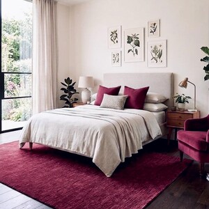 Puede incluir: Un dormitorio con una cama grande con cabecero tapizado en beige, ropa de cama beige claro y dos cojines de color rojo intenso. Una alfombra de color rojo intenso cubre el suelo de madera oscura y un sill&oacute;n de terciopelo rojo intenso a la derecha.