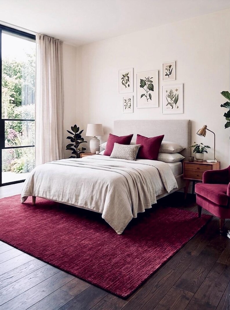 Puede incluir: Un dormitorio con una cama grande con cabecero tapizado en beige, ropa de cama beige claro y dos cojines de color rojo intenso. Una alfombra de color rojo intenso cubre el suelo de madera oscura y un sill&oacute;n de terciopelo rojo intenso a la derecha.