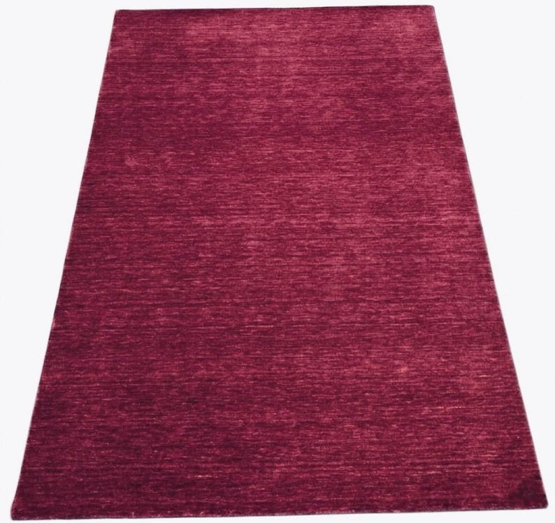 Puede incluir: Una alfombra rectangular de color magenta intenso y s&oacute;lido. La superficie presenta un sutil patr&oacute;n de tejido texturizado horizontal que crea ligeras variaciones en la intensidad del tono magenta a lo largo de toda la alfombra.