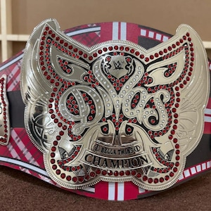 May include: A championship belt with a silver and red design. The belt features the words "BELLA TWINS CHAMPION" and the WWE logo. The main plate has red gemstones and a wing design. The belt's strap has a red and black plaid pattern.