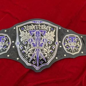 Puede incluir: Un cinturón de campeonato de lucha libre negro y plateado con detalles morados. El cinturón presenta el nombre "Undertaker" y la frase "The Phenom". Las placas laterales enumeran "The Deadman", "The Last Ride", "American Badass" y "Big Evil".