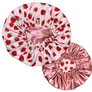 May include: Two hair bonnets featuring a light pink fabric with a repeating red strawberry pattern. One bonnet shows the strawberry print on the exterior and a solid pink satin interior, while the other shows a solid pink satin exterior and strawberry print interior.