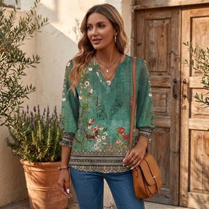 May include: A green three-quarter sleeve blouse featuring a floral print with red and white flowers, a V-neckline, and a decorative patterned border at the hem. The blouse is paired with blue denim jeans and a brown leather shoulder bag.