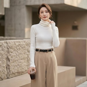 May include: A white ribbed turtleneck sweater with long sleeves paired with high-waisted tan trousers and a thin black belt with a gold buckle. The sweater features a fitted silhouette and a high neck, while the trousers have a tailored cut.