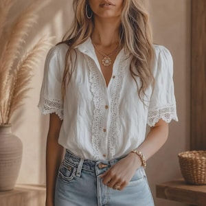 May include: A white short-sleeved button-down shirt featuring intricate lace trim along the front placket and sleeve cuffs. The shirt is paired with light blue denim jeans. A gold pendant necklace and a gold beaded bracelet are worn as accessories.