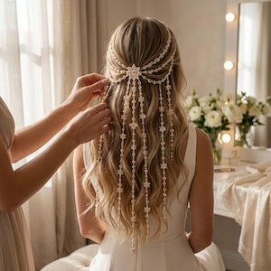 Puede incluir: Un accesorio para el cabello de color crema con un motivo central en forma de estrella y múltiples hilos en cascada adornados con pequeñas cuentas en forma de flor, dispuestos sobre el cabello largo y ondulado con un efecto decorativo.