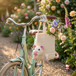 Könnte beinhalten: Eine cremefarbene Häkeltasche in Form einer Maus mit rosa Ohren, einer rosa Nase und einem rosa Schwanz, die am Lenker eines hellgrünen Fahrrads hängt. Die Tasche enthält einen Blumenstrauß aus rosa Rosen und violetten Blüten.