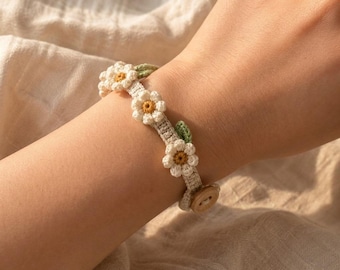 Puede incluir: Una pulsera de ganchillo con tres flores blancas de centros amarillos, acentuadas por pequeñas hojas verdes. La banda está hecha de hilo color crema y se asegura con un cierre de botón de madera, creando un diseño floral en la muñeca.