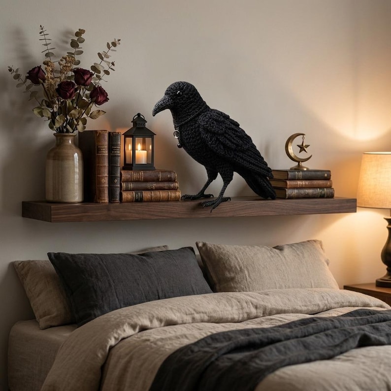 May include: A black crocheted raven figurine with a small silver pendant around its neck, positioned on a wooden shelf alongside a vase of dried roses, several vintage books, a small black lantern with a candle, and a gold crescent moon ornament.