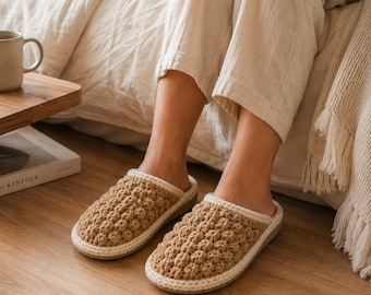 Puede incluir: Un par de zapatillas de ganchillo color canela con un patrón texturizado de puntos de burbuja en la parte superior. Las zapatillas tienen un borde sólido de color crema claro alrededor de la abertura y una suela plana, tipo calzado.