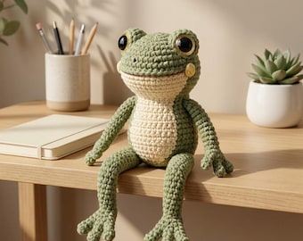 Puede incluir: Un peluche de rana tejido a ganchillo con cuerpo verde salvia, vientre color crema y grandes ojos negros con iris dorados. El animal de peluche tiene extremidades largas y articuladas y está sentado erguido con las piernas colgando del borde.