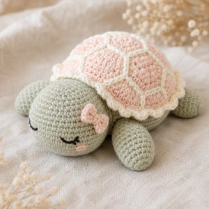 May include: A crocheted turtle toy with a sage green body and a pale pink shell featuring cream-colored stitching. The turtle has embroidered closed eyes, a small pink blush on its cheek, and a tiny pink bow attached to the side of its head.