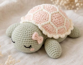 Puede incluir: Un juguete de tortuga tejido a ganchillo con cuerpo verde salvia y caparazón rosa pálido con costuras color crema. La tortuga tiene ojos bordados cerrados, un pequeño rubor rosa en la mejilla y un diminuto lazo rosa en la cabeza.