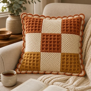 May include: A square crochet throw pillow featuring a checkered pattern of waffle-stitch squares in burnt orange, cream, and mustard yellow. The pillow is finished with a decorative scalloped cream-colored border along all four edges.