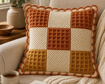 Puede incluir: Un cojín decorativo cuadrado de ganchillo con un patrón de cuadros de punto de gofre en tonos naranja quemado, crema y amarillo mostaza. El cojín está rematado con un borde festoneado de color crema en los cuatro lados.