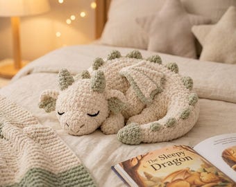 Puede incluir: Un peluche de dragón tejido a ganchillo en color crema con detalles en verde salvia en los cuernos, las alas y las puntas de la cola. El dragón aparece en posición de reposo con los ojos cerrados, con una textura suave y cola enroscada.