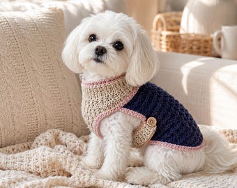 Puede incluir: Un perro pequeño de color blanco lleva puesto un suéter de ganchillo con un panel frontal beige, una parte trasera azul marino oscuro y un borde rosa claro. La prenda incluye un pequeño bolsillo beige en el lateral con un botón de madera.