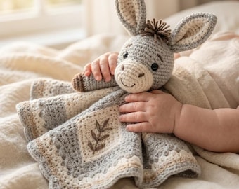 May include: A crochet security blanket featuring a grey donkey head with long ears and a small tuft of brown hair. The attached blanket is square-shaped with a cream-coloured centre panel displaying a brown embroidered leaf design and a grey border.