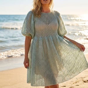 Könnte beinhalten: Ein helles, salbeigrünes Häkelkleid mit rundem Ausschnitt, kurzen Puffärmeln und einem ausgestellten Rock. Das Kleidungsstück ist in einem durchbrochenen Strickmuster gefertigt, das eine halbtransparente Textur erzeugt und über dem Knie endet.