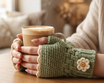 Puede incluir: Un par de guantes sin dedos de color verde salvia con un patrón de tejido de cesta texturizado, un borde festoneado color crema y una pequeña flor de ganchillo blanca con centro amarillo en el dorso de cada mano, sosteniendo una taza.