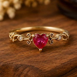 Op de afbeelding: Een gouden ring met een centrale hartvormige rode edelsteen. De band is voorzien van gedetailleerd krulwerk aan beide zijden van het hart, geaccentueerd door kleine, heldere, rondgeslepen steentjes in de metalen versieringen op de band.