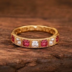 May include: A gold band ring featuring an alternating pattern of square-cut red rubies and clear white stones set in a beaded metal frame. The ring displays the text PRIMEJELLERYUS faintly in the background, showcasing a repeating gemstone design.