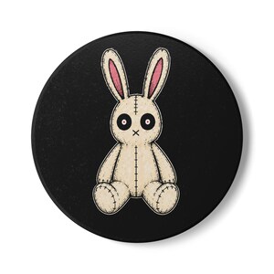 Op de afbeelding: Een ronde zwarte onderzetter met een gecentreerde illustratie van een beige knuffelkonijn. Het konijn heeft lange oren met roze binnendelen, grote zwarte ronde ogen met rode pupillen, een gestikte mond en zichtbare donkere naadlijnen.