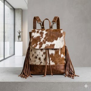 May include: A brown and white cowhide backpack featuring a fold-over flap, a drawstring closure, and long leather fringe detailing on both sides. The bag includes adjustable shoulder straps with metal hardware and a top handle for carrying.