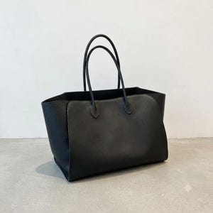 May include: A black tote bag with a textured surface, featuring two tall, slender handles attached to the top edge. The bag has a wide, open-top design with a structured base and curved side panels that extend upward to meet the handle attachments.