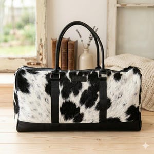 May include: A rectangular duffel bag featuring a black and white cow print pattern with a textured hair-on-hide finish. The bag includes black leather-like trim at the base, black nylon webbing handles, and a small rectangular brand label on the front.